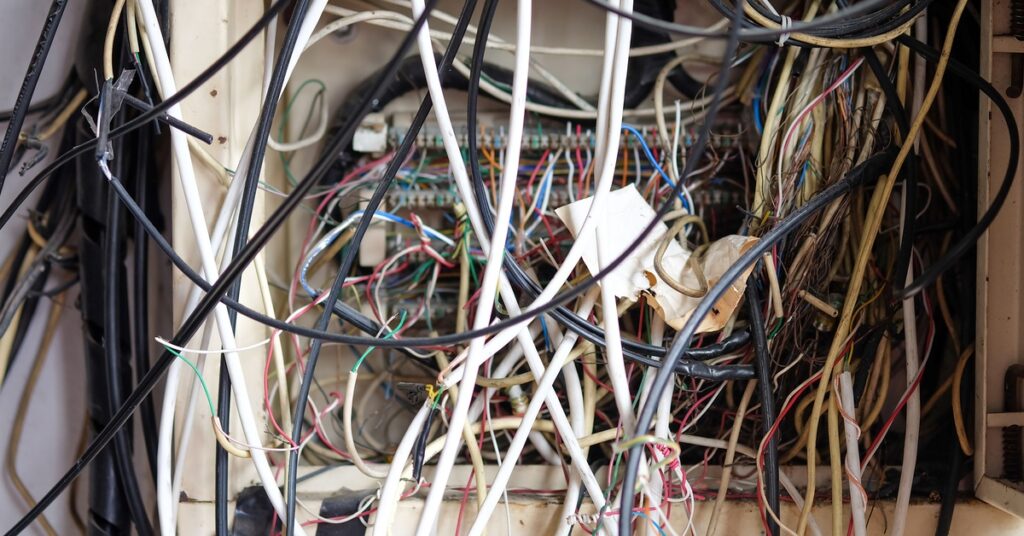 An electrical fuse box is completely open. Several cables in assorted colors are messily tangled up inside.
