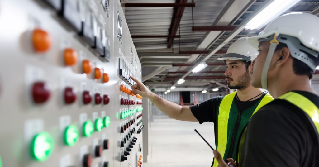 Two men in hard hats and hi-vis vests stand in front of a large electrical panel with several buttons. One man points at it.