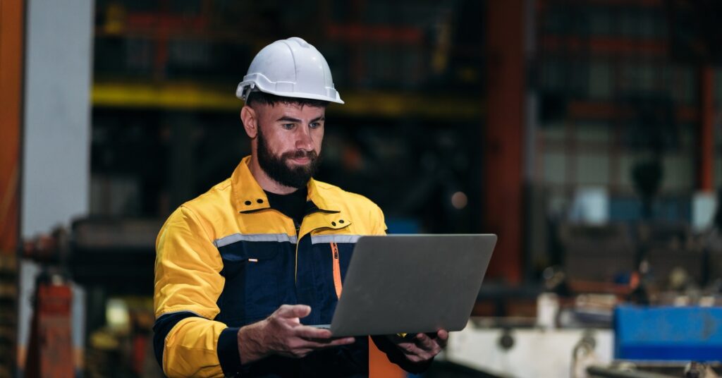 A man in a hard hat and yellow jacket stands in a darkened warehouse. He holds a laptop and looks at the screen.