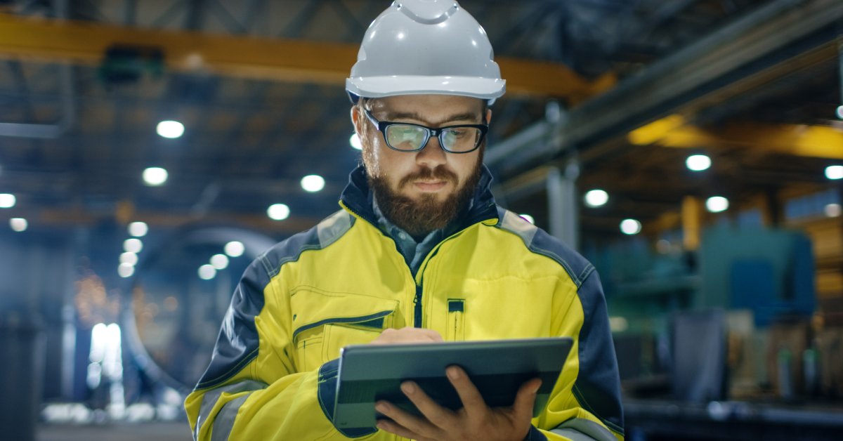 A man in a hard hat and yellow jacket stands in an empty warehouse. He glances at the screen of his tablet.