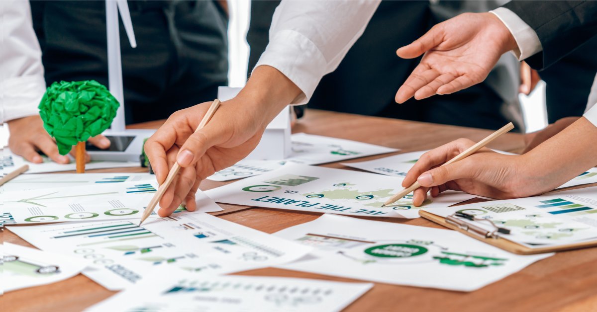 Several people stand in front of a table strewn with paperwork about sustainability. They point and gesture at various papers.