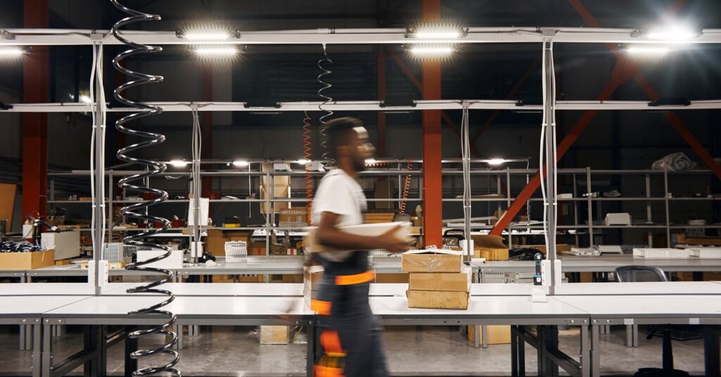 a warehouse lit by bright LED lights. A worker in a black apron walks quickly while carrying a box.