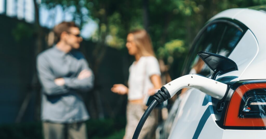 In the foreground, a charger is plugged into an electric car. In the background, a man and a woman chat.